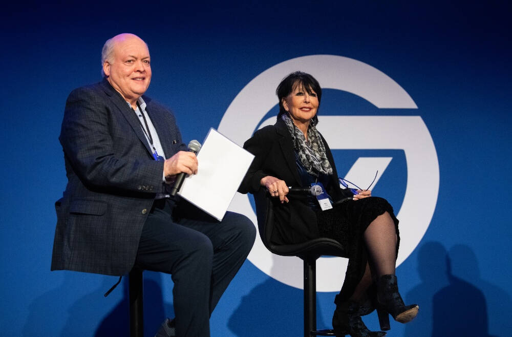 Two people are seated on stage in front of a blue backdrop with a GVSU logo. The person on the left, wearing a dark blazer and blue pants and holding a microphone and papers. The person on the right, wearing a black blazer, heeled boots.